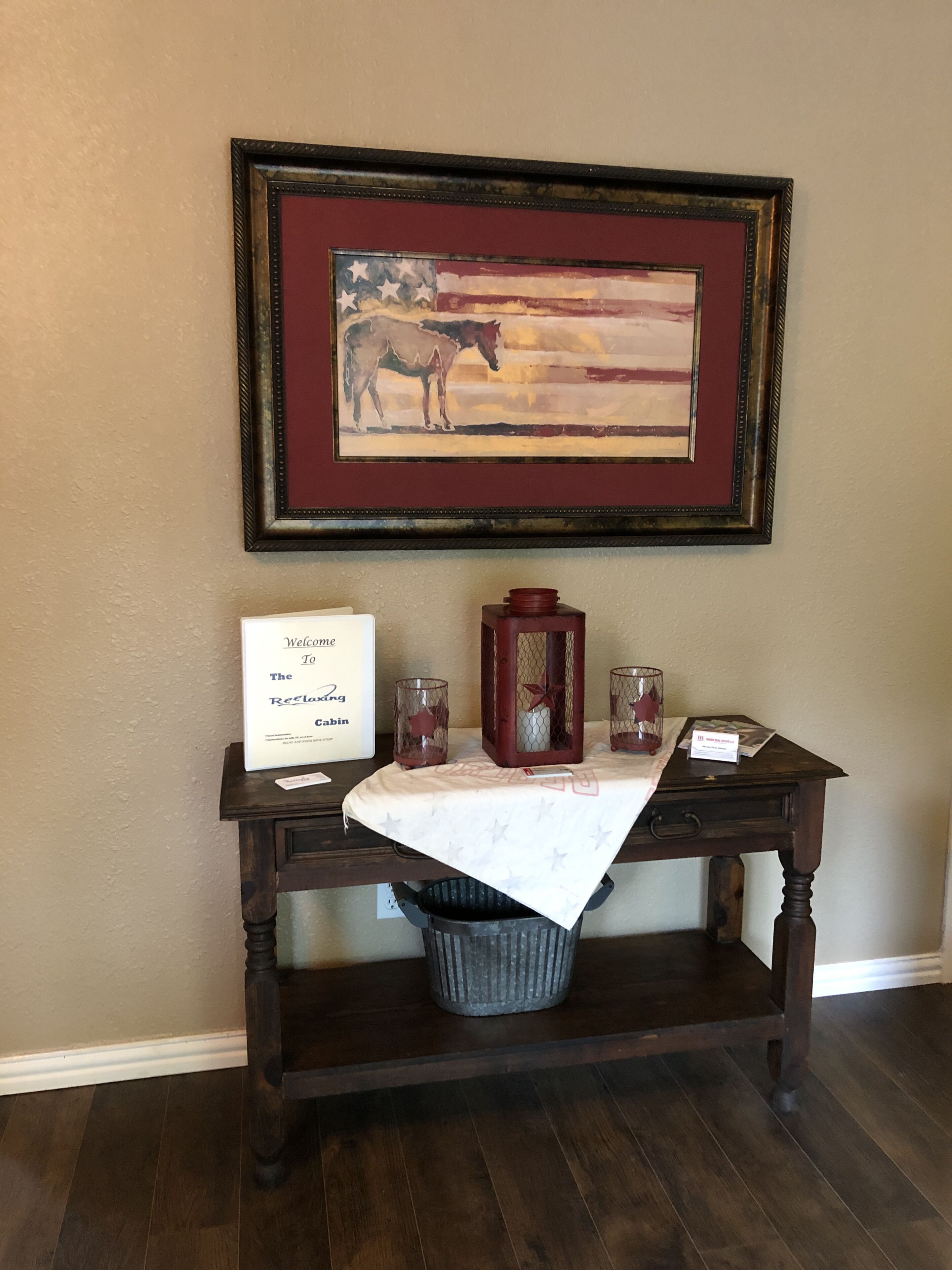 A framed painting of a horse on a wall, a welcome sign, a candle holder, and a small table with a tablecloth.