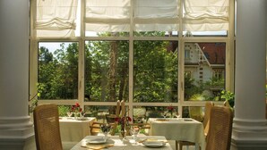 A dining area with a table set for two, large windows with curtains, and a view of greenery and buildings outside. - Charmant appartement entièrement équipé (ARCACHON)