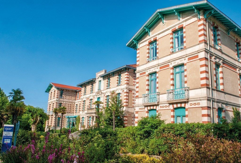 Ein historisches Gebäude mit einer rot-weiß gestreiften Fassade, mehreren Balkonen und üppigem Grün im Vordergrund.
