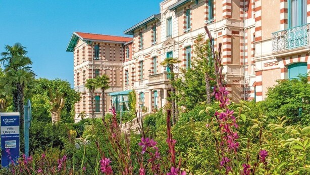 A large, historic building with a red-tiled roof and striped facade, surrounded by lush greenery and flowering plants. - Charmant appartement entièrement équipé (ARCACHON)