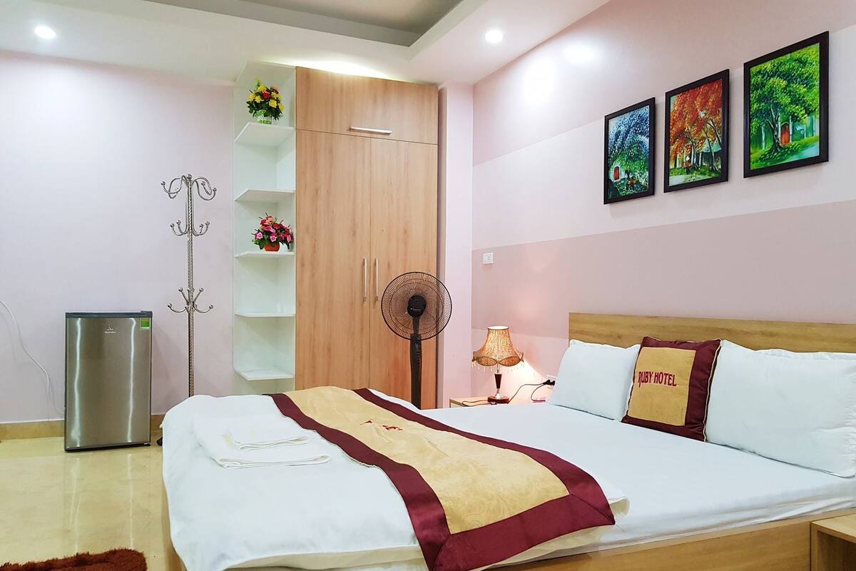 A hotel room with a bed, a wardrobe, a fan, a lamp, and a refrigerator.