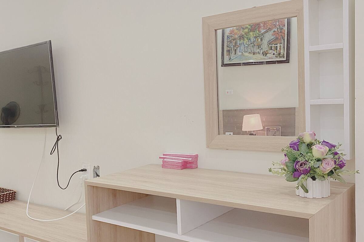 A wooden desk with a flat-screen TV mounted above it, a small pink box, and a vase with flowers on the right side.