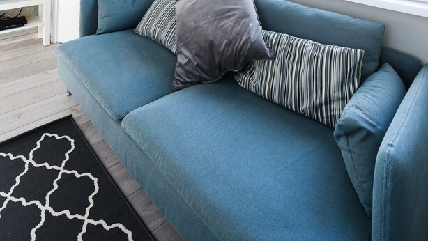 A modern living room with a blue sofa, a television on a white stand, and a large window with a view of buildings and trees. - Apartment Lali - Apartment with Balcony (Zadar)