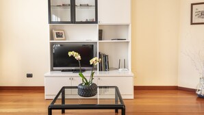Ein Wohnzimmer mit Flachbildfernseher, weißem Unterhaltungsmöbel und einem Glastisch mit Pflanzgefäß im Reifen-Design.