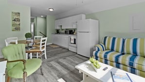 A modern kitchen with white cabinets, a refrigerator, and a dining area with a green upholstered chair and a wooden table. - "Sand Dollar Suites" suite 6: A short stroll from our sandy shores! (Carolina Beach)