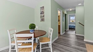 A dining area with a round table, white chairs, and a potted plant. A hallway leads to a bedroom with a bed and a window. - "Sand Dollar Suites" suite 6: A short stroll from our sandy shores! (Carolina Beach)