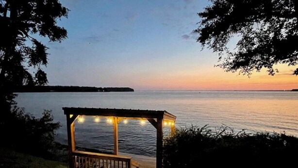 A wooden deck with a pergola and string lights overlooking a calm body of water at dusk. - Nancy’s Touch, A Waterfront Paradise! (North East)