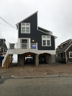 A two-story house with a balcony, a covered driveway, and a wooden staircase leading to the entrance. - Oceanfront/ Beachfront Home on Sand Hills Scituate (Scituate)