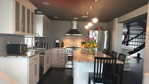 Une cuisine moderne avec des meubles blancs, un îlot central, des appareils électroménagers en acier inoxydable et un escalier noir menant à une mezzanine.