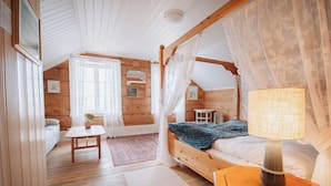 Un dormitorio acogedor con una cama con dosel de madera, una mesita de noche con lámpara, una pequeña mesa con un jarrón y una ventana con cortinas translúcidas.