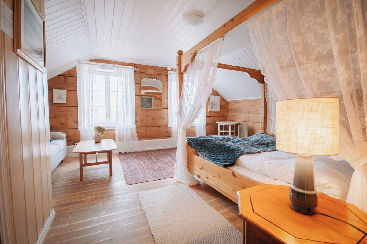 Un dormitorio acogedor con una cama con dosel de madera, una mesita de noche con lámpara, una pequeña mesa con un jarrón y una ventana con cortinas translúcidas.