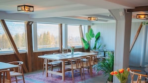 Un comedor con mesas y sillas de madera, un ventanal con vistas y una planta con flores anaranjadas.