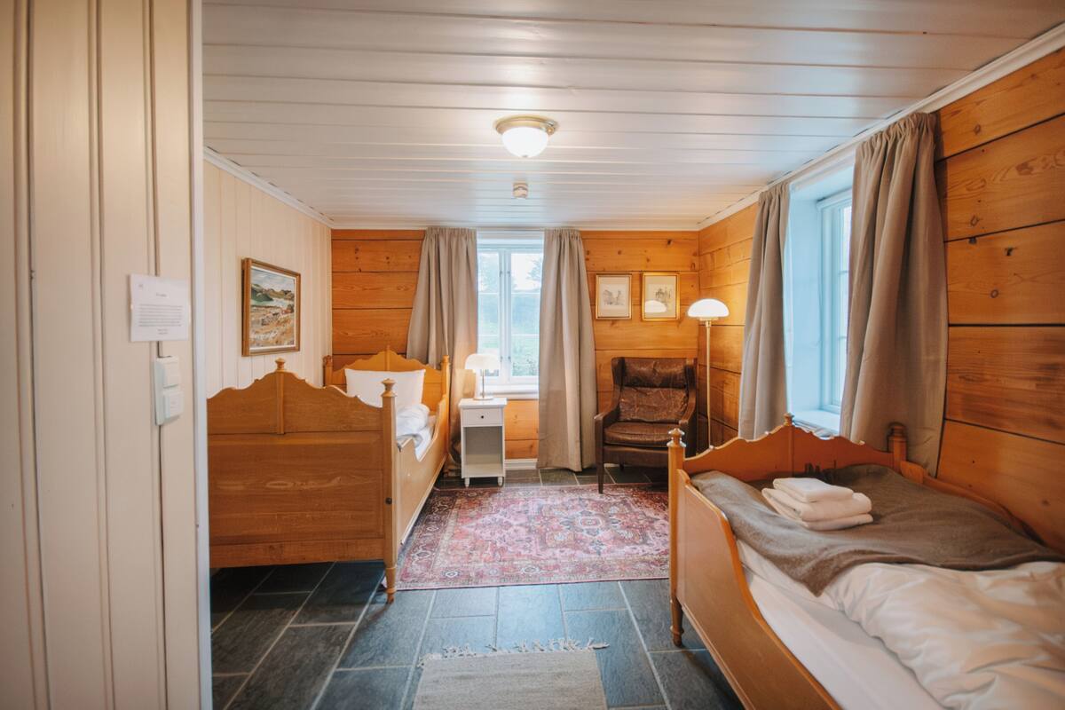 Una habitación con paneles de madera, dos camas, una mesita de noche, una silla y un ventanal con cortinas.