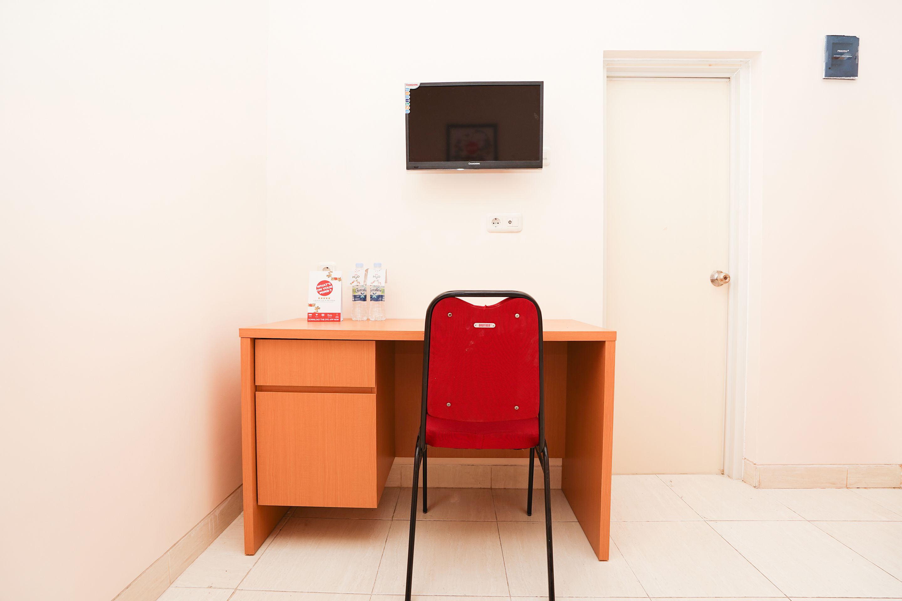 A small, clean room with a wooden desk, a red chair, a flat-screen TV mounted on the wall, and a door with a light switch.