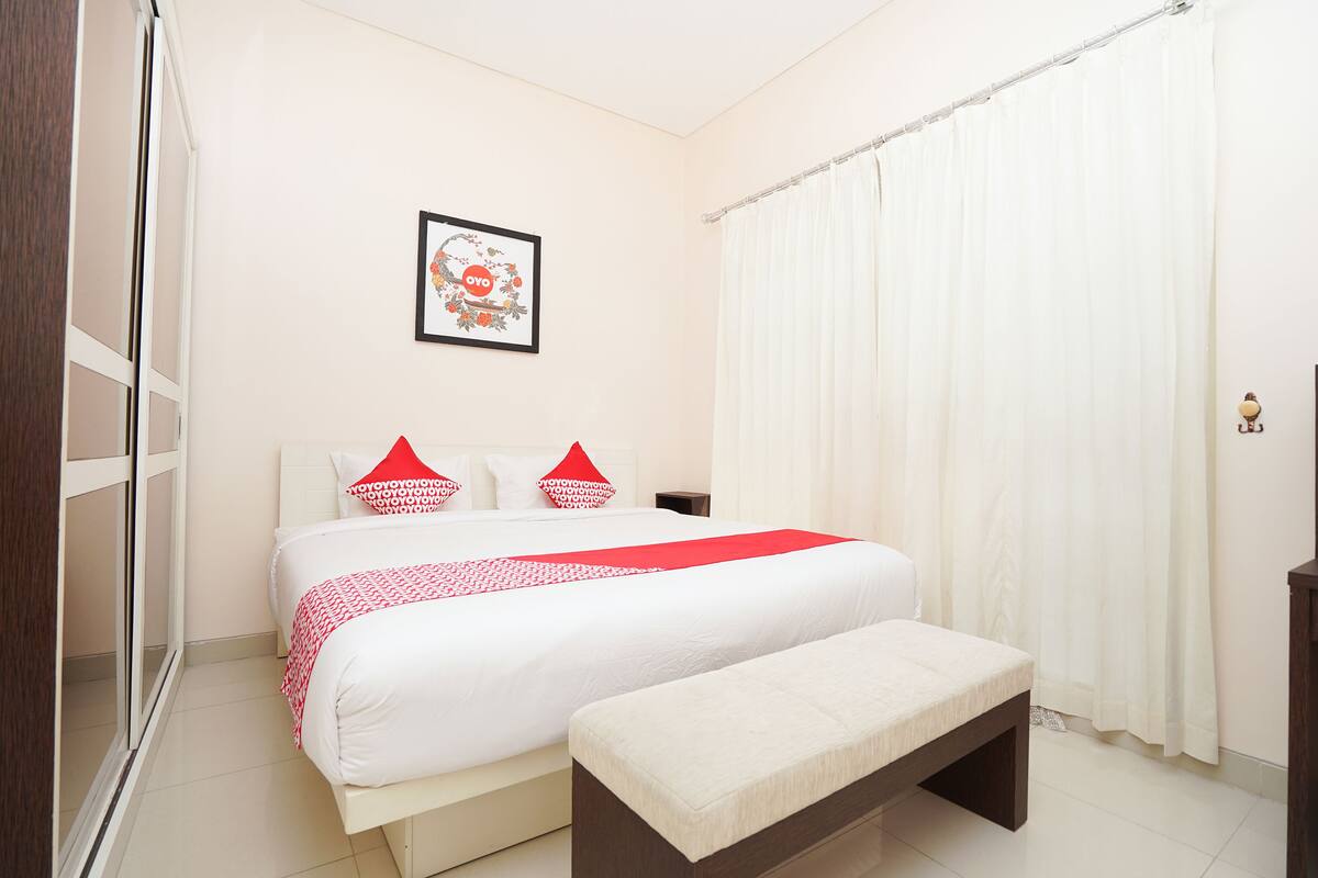 Une chambre à coucher avec un lit, une banquette, une table de chevet, un tableau encadré au mur et un rideau.