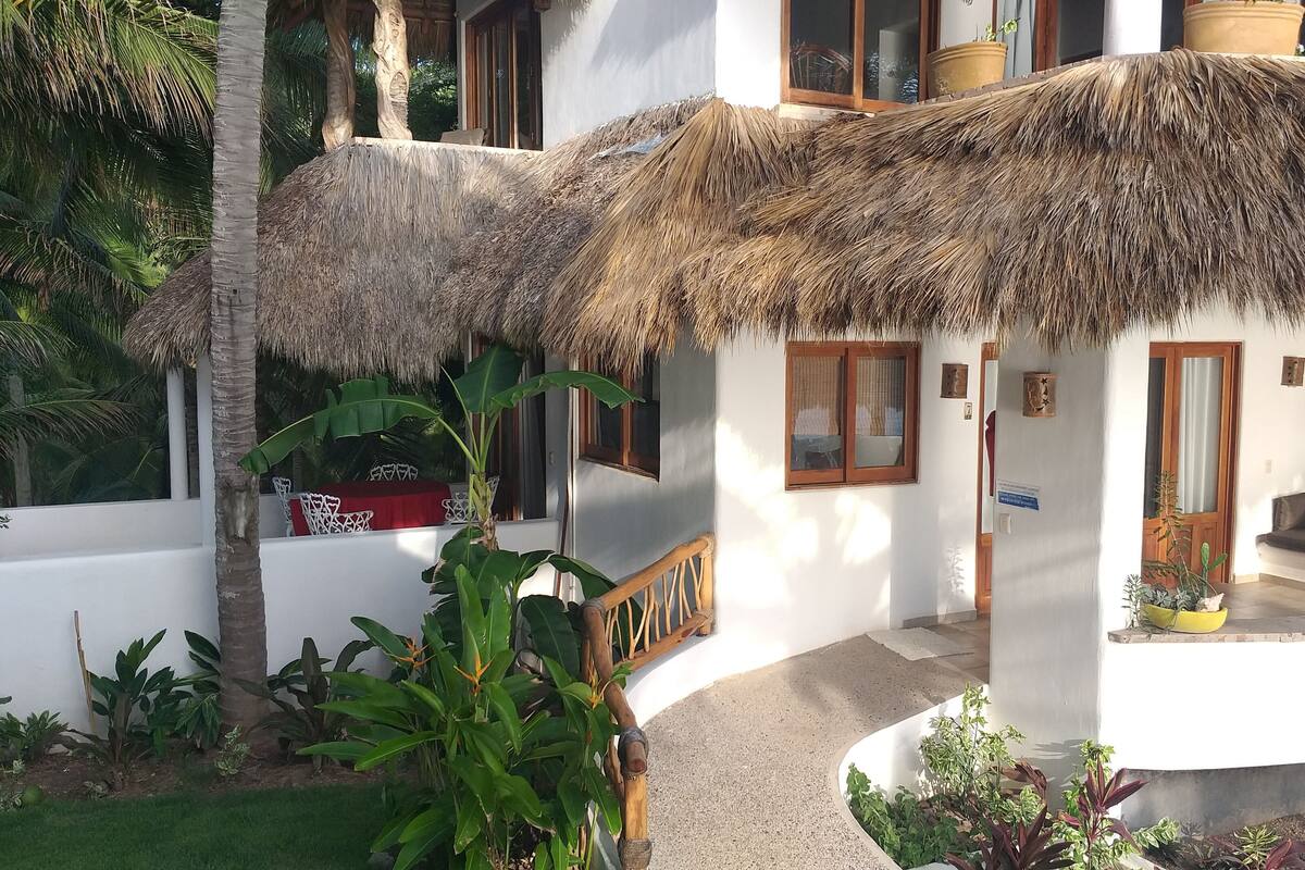 A white building with a thatched roof, a wooden staircase, and a seating area with cushions.