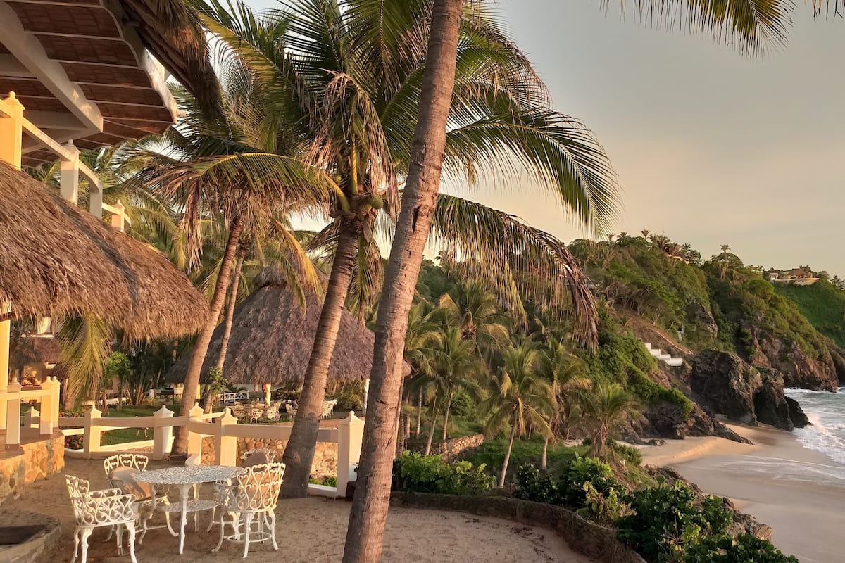 A beachfront property with thatched-roof structures, palm trees, and a sandy beach.