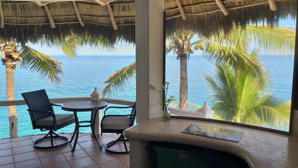 A balcony with a table and chairs, overlooking a beach with palm trees and the ocean.