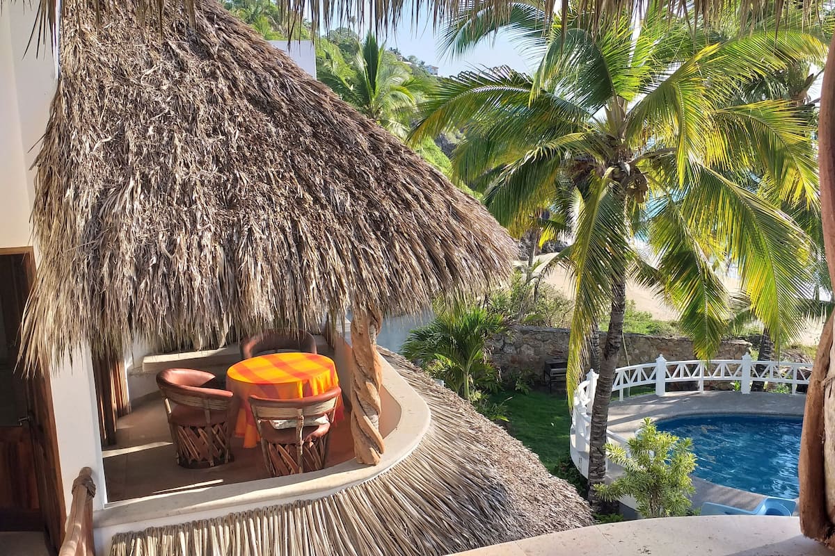 A thatched-roof outdoor seating area with a pool and palm trees.