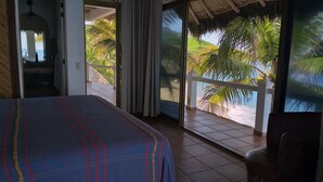 A bedroom with a bed, a chair, and a view of a beach and palm trees through a sliding glass door. - Bungalows Lydia (San Francisco)