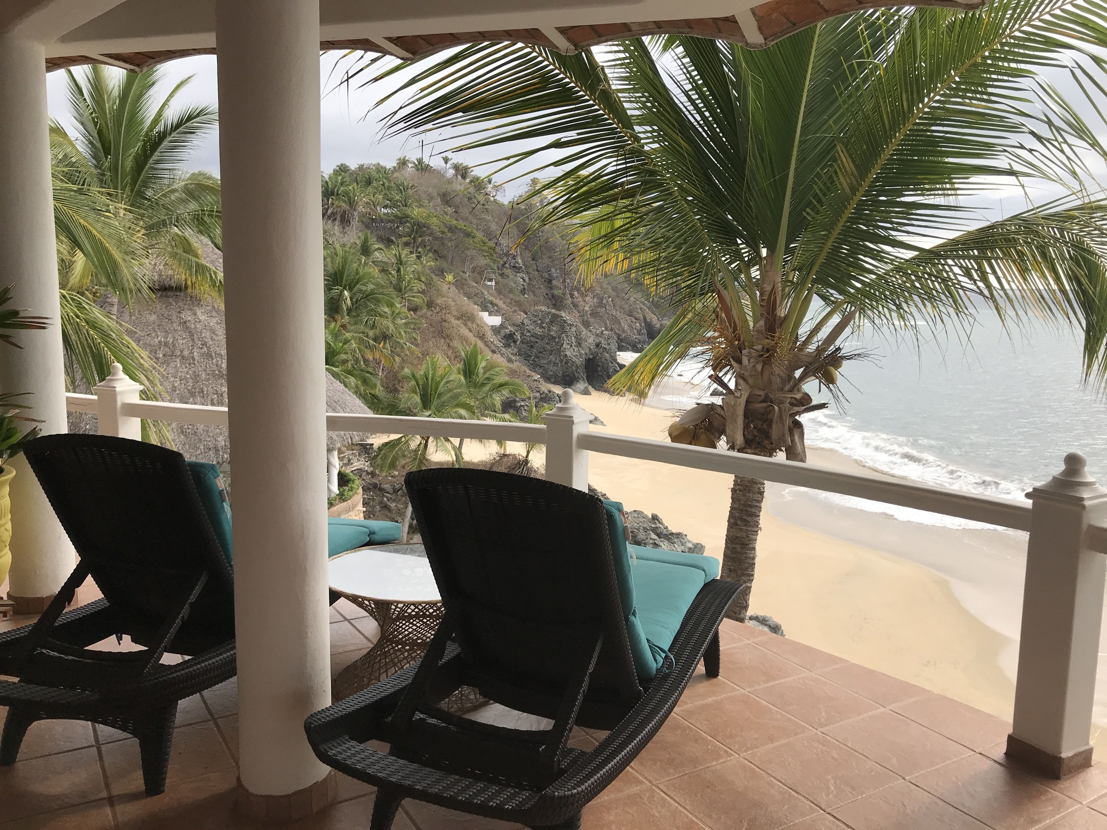 A covered outdoor seating area with wicker chairs and a table, overlooking a beach with palm trees and a rocky hillside.