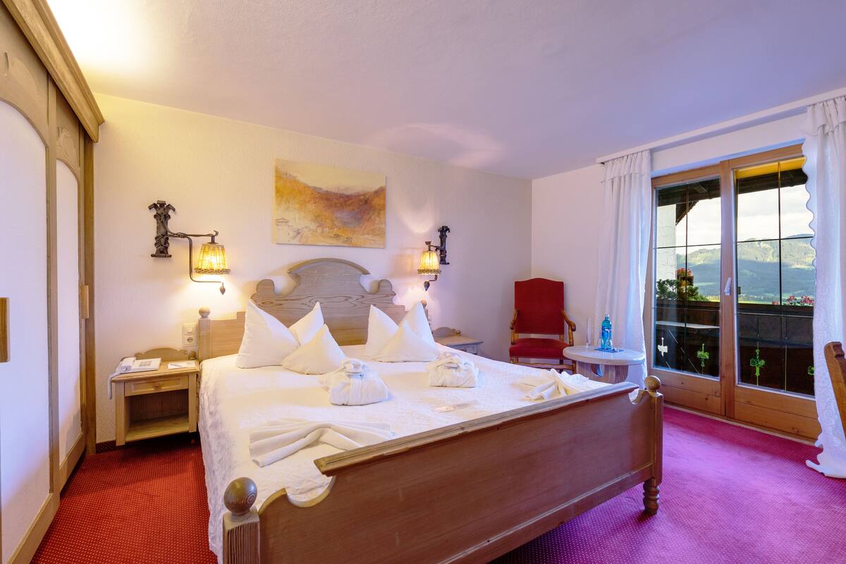 Una habitación de hotel con una cama de madera, una silla roja, una mesita y un balcón con vistas.