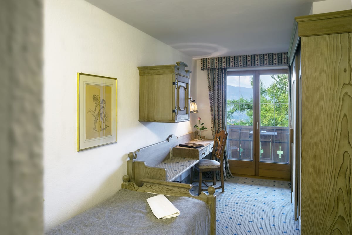 Una habitación con una cama, un escritorio, una silla, un ventanal con vistas y una puerta que da a un balcón.