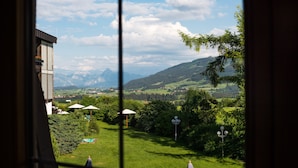 Vista a través de una ventana de un paisaje verde exuberante con montañas al fondo, un edificio con balcón y un farol.