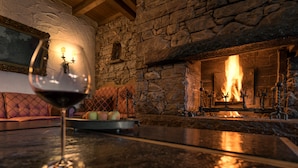 Una chimenea de piedra con fuego, una copa de vino sobre una mesa y un banco acolchado.