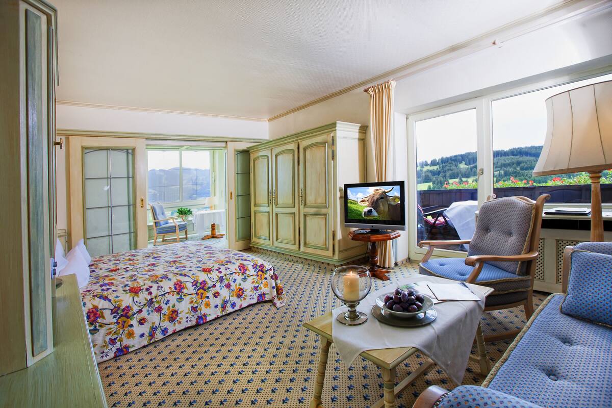 Habitación de hotel con una cama, televisión, una mesa con vela y fruta, y vistas a un bosque a través de amplias ventanas.