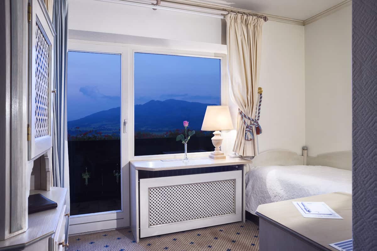 Una habitación de hotel con una cama, una mesita de noche con lámpara, una ventana que da a vistas de montañas y una puerta con un enrejado decorativo.