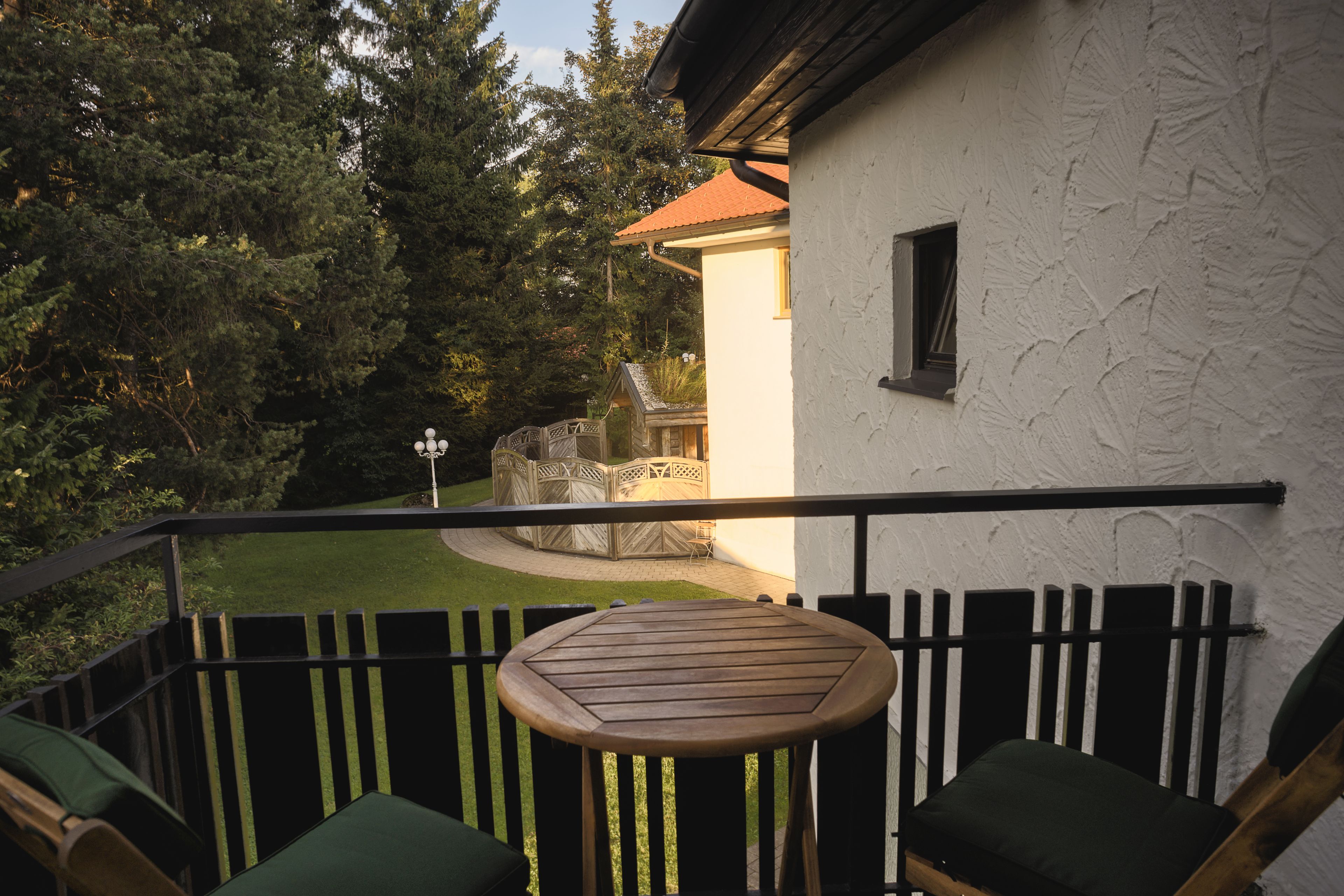 A balcony with a wooden table and chairs, a white building with a red roof, and a garden area with trees and a lawn.