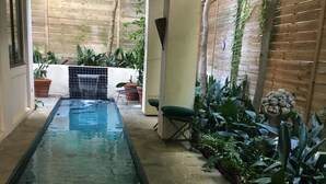 An outdoor pool area with two green lounge chairs, a small table, and a stone bench. - Artist’s Library; Private Swimming Pool in River Oaks Area (Houston)