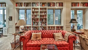 A living room with a red leather sofa, a bookshelf filled with books, and a window with a view of trees. - Artist’s Library; Private Swimming Pool in River Oaks Area (Houston)