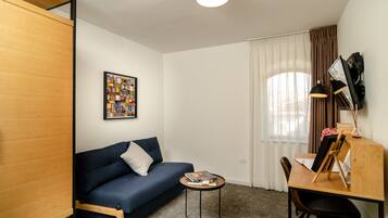 A small, modern room with a blue sofa, a wooden desk with a chair, a small round table, and a large window with curtains.