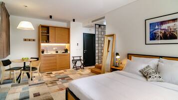 A hotel room with a bed, a desk with a chair, a small table with a bowl of fruit, a coffee machine, and a kitchen area with cabinets and a sink.