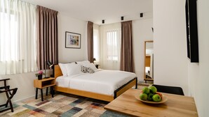 Habitación de hotel con una cama, una mesita de noche con lámpara, una pequeña mesa con un frutero y vista al pasillo.