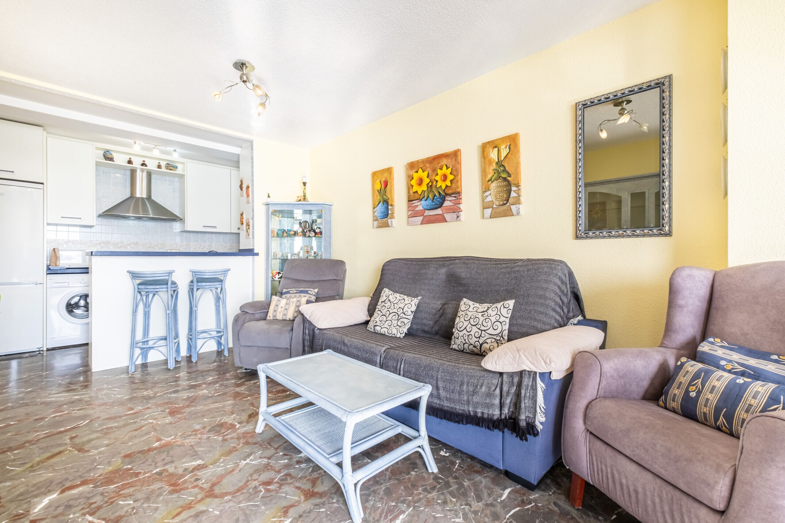 A living room with a grey sofa, a coffee table, and a kitchen in the background.
