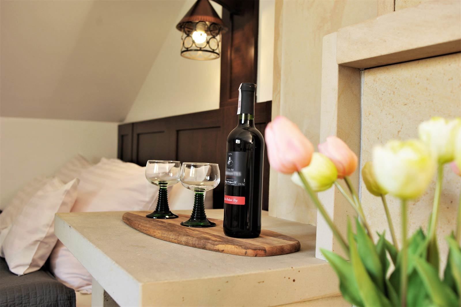 A bedroom with a wooden headboard, a bedside table with a bottle of wine and two glasses, and a flower arrangement.