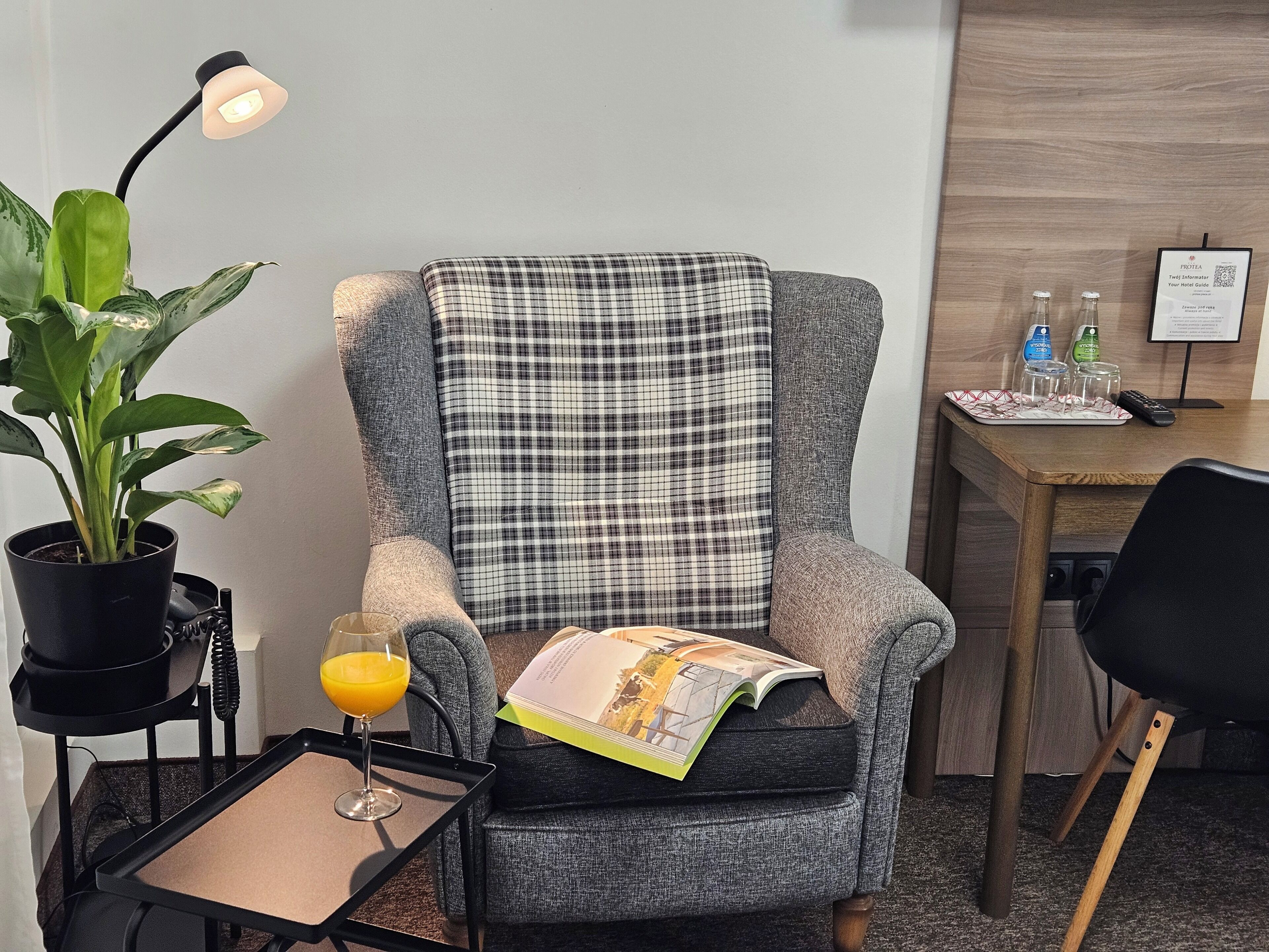 Una habitación de hotel moderna con un sillón gris, una mesita con un vaso de zumo de naranja, una planta en maceta y una lámpara.