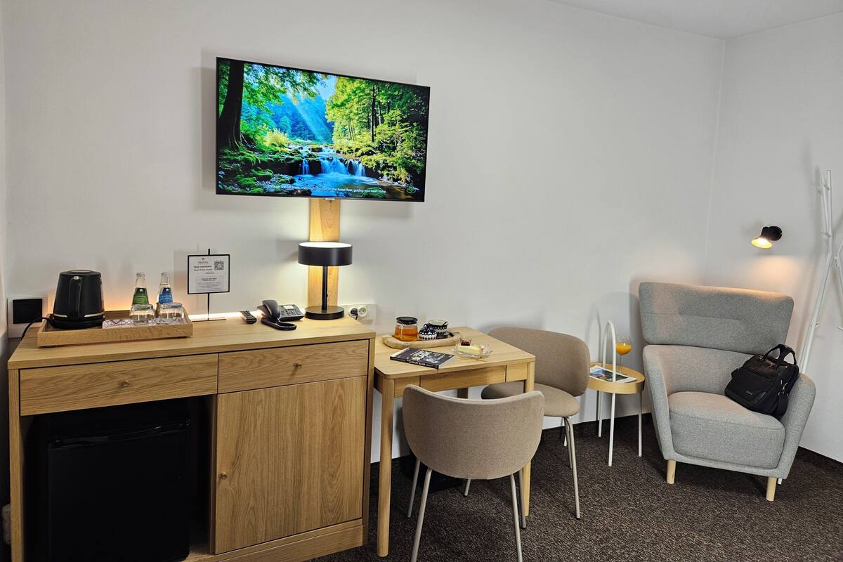A hotel room with a TV displaying a nature scene, a desk with a lamp, a chair, a small table, and a bedside table with a lamp.