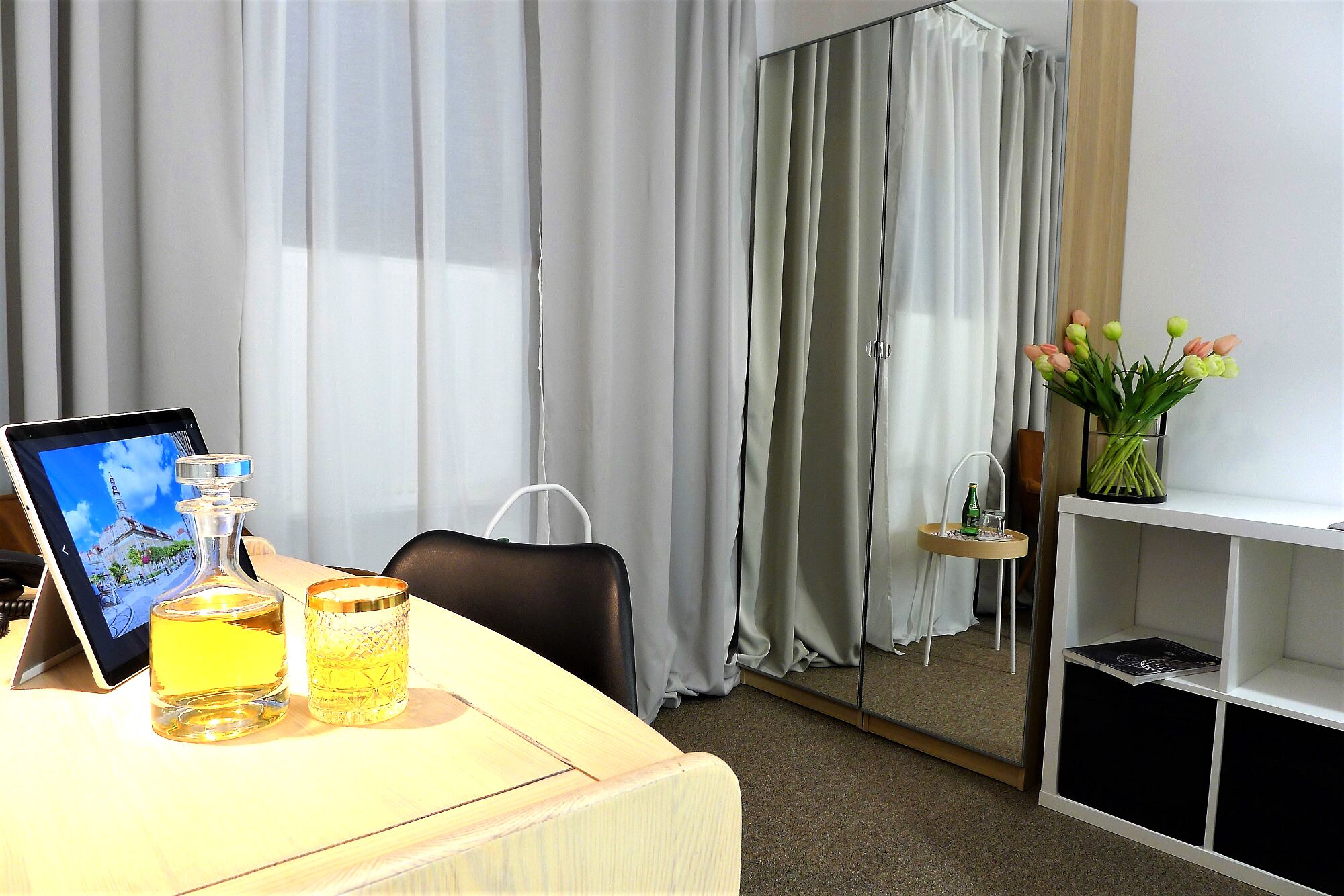 A hotel room with a desk, a chair, a bedside table with a lamp, a mirror, and a shelf with flowers.