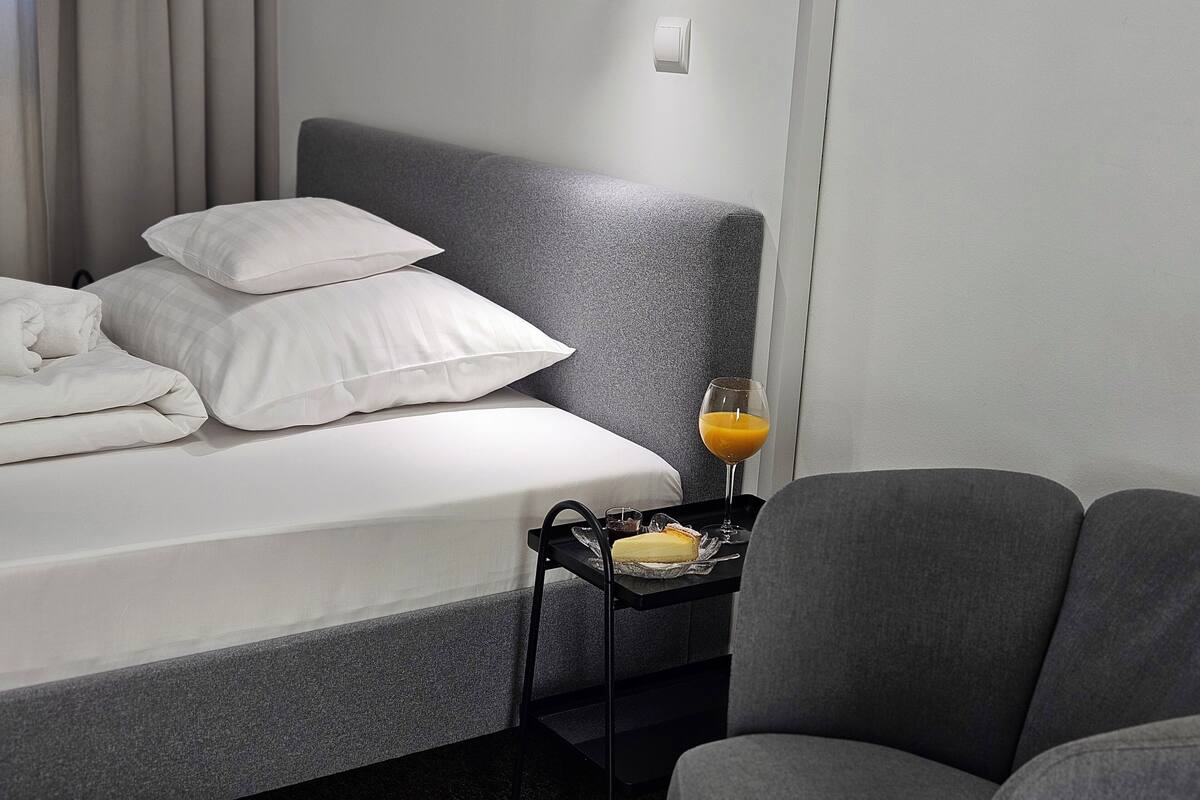 A neatly made bed with pillows, a bedside table with a glass of orange juice, and a chair.