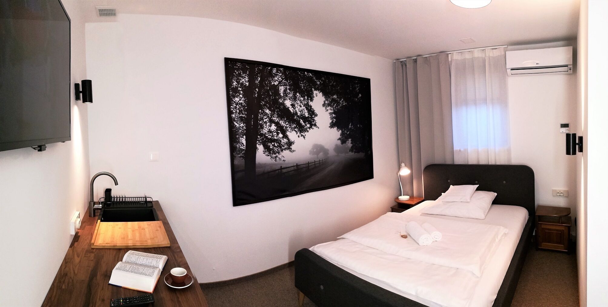 A hotel room with a bed, bedside table, lamp, and a painting of a forest scene.