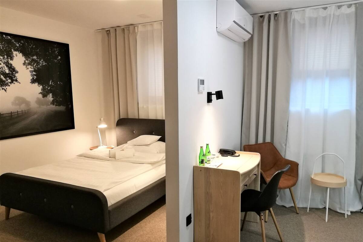A hotel room with a bed, desk, chair, and a painting of a landscape.