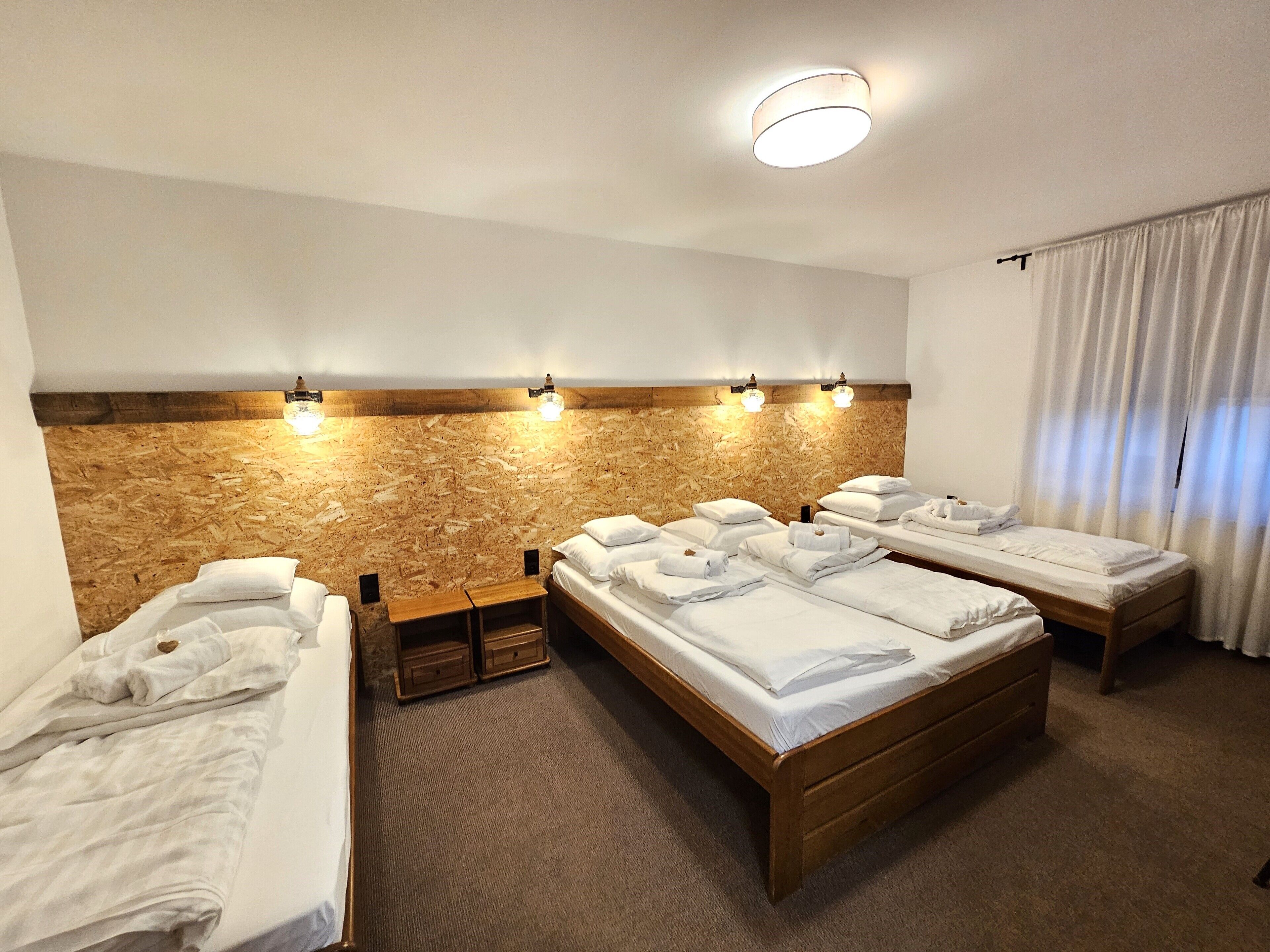 Una habitación de hotel con cuatro camas individuales, cabecera de madera, lámpara de pared y ventana con cortinas.