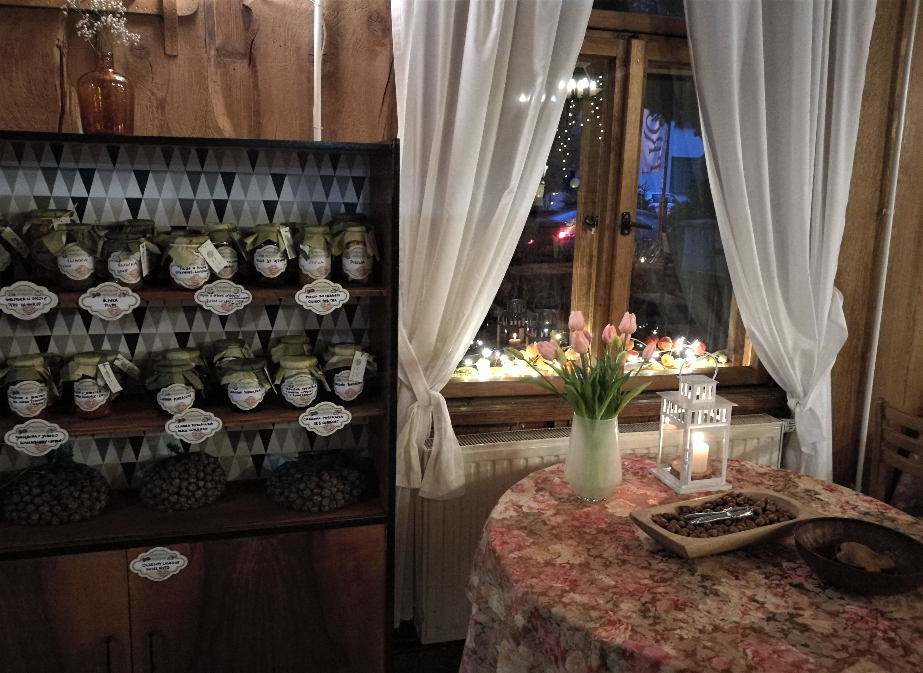A room with a wooden shelf displaying jars of preserves, a table with a floral tablecloth, candles, and a vase with flowers.