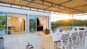 Un espace extérieur aménagé pour les repas, avec une table et des chaises en bois, une porte d’entrée en verre et une vue sur le paysage.
