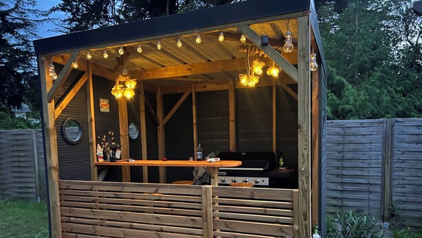 A wooden outdoor bar with a grill, hanging light bulbs, and a wooden fence in the background. - ILE TUDY - Contemporary house 6 places with garden 100m from the beach (Île-Tudy)