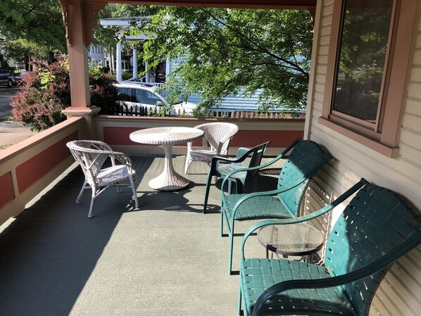 A covered outdoor seating area with wicker chairs and tables, a green sofa, and a view of parked cars and buildings in the background.